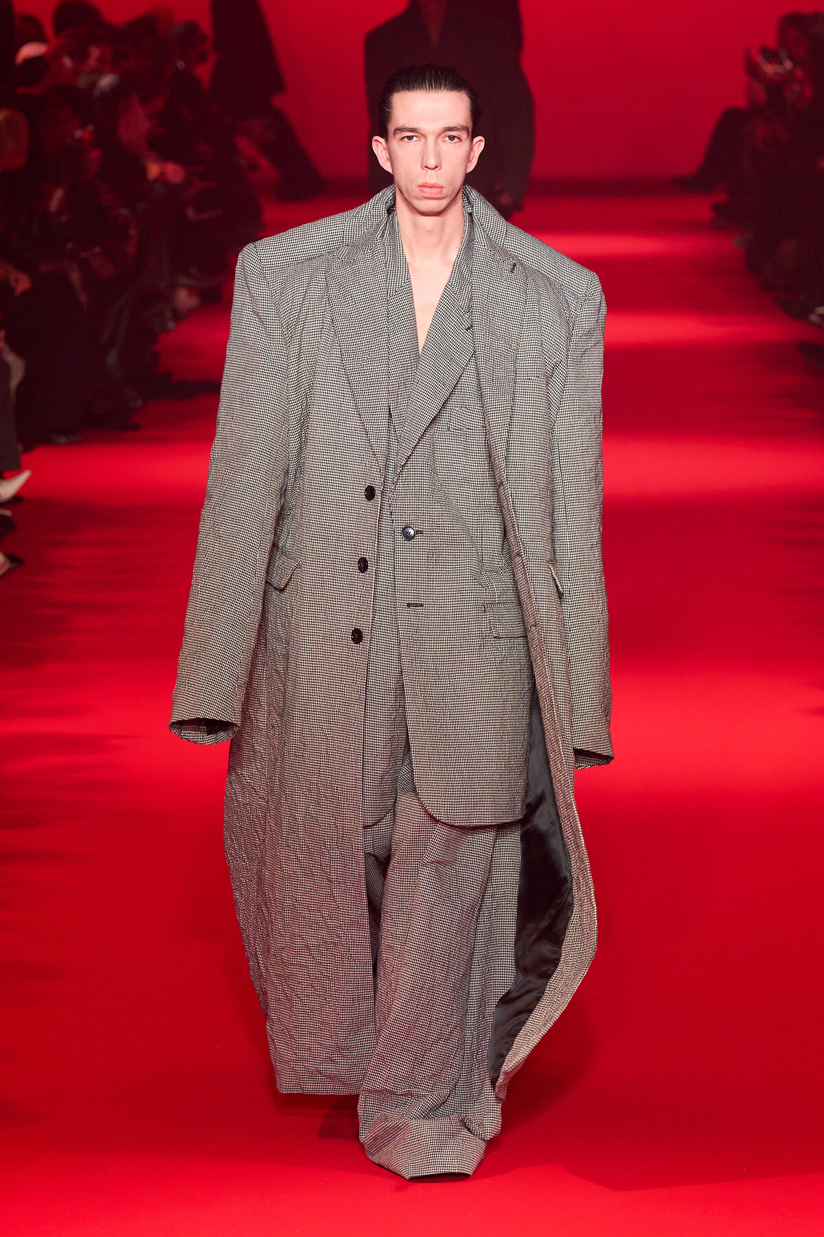 Male model wearing an oversized gray checked suit walking a red runway.