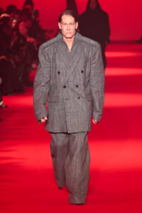 Male model walks a runway in an oversized gray plaid double-breasted suit against a red backdrop.