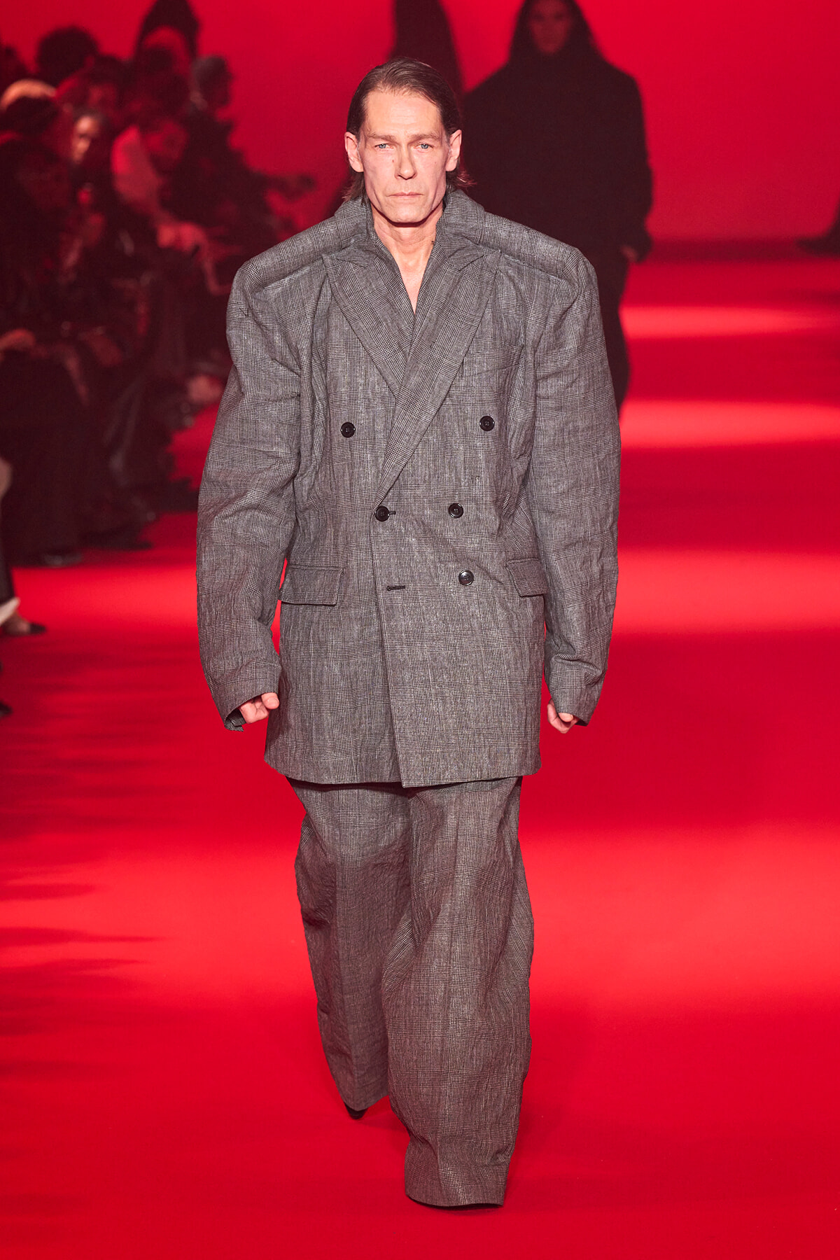Male model walks a runway in an oversized gray plaid double-breasted suit against a red backdrop.