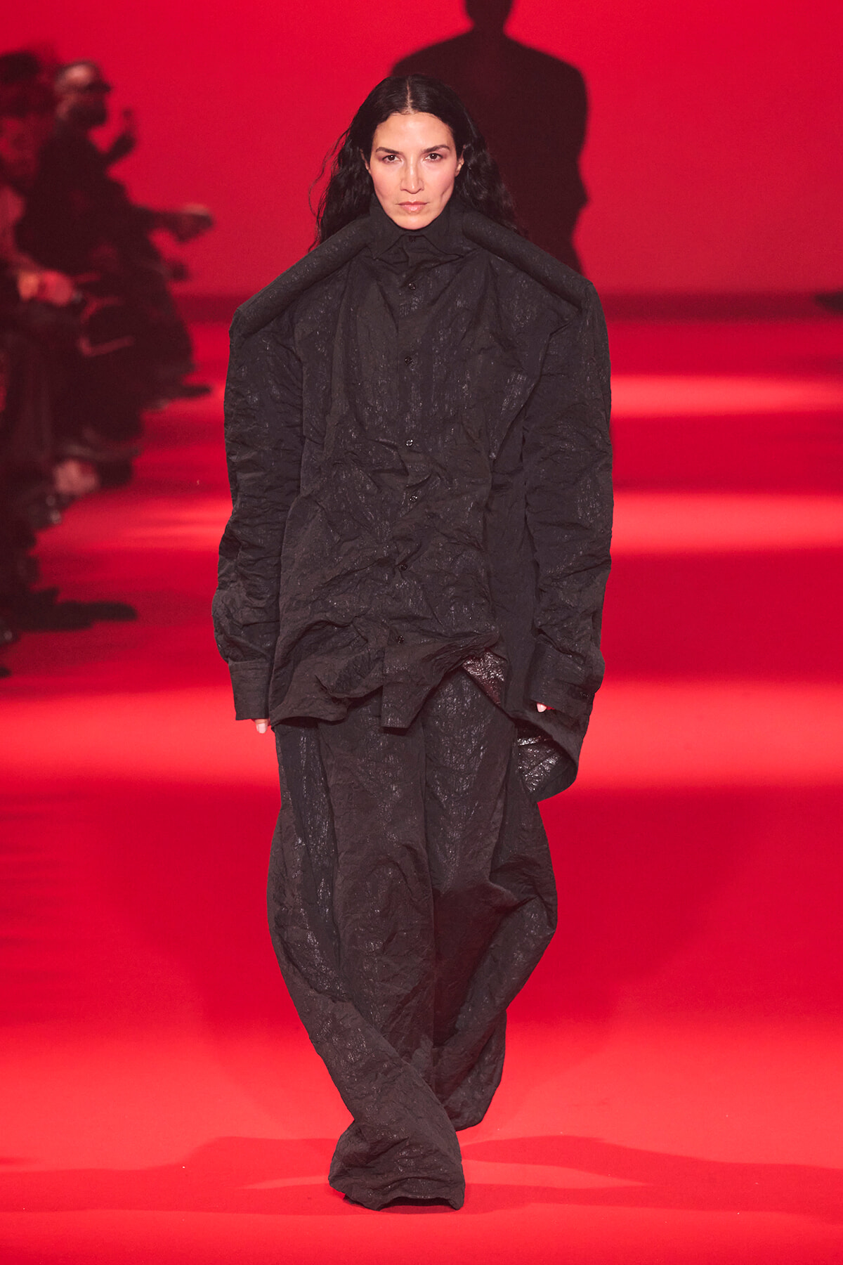 Model walking the runway in an oversized crinkled black outfit against a red backdrop on a fashion show.