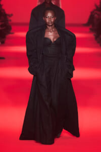 Female model walking a fashion show runway in a black corset top and long black coat against a red-lit backdrop.