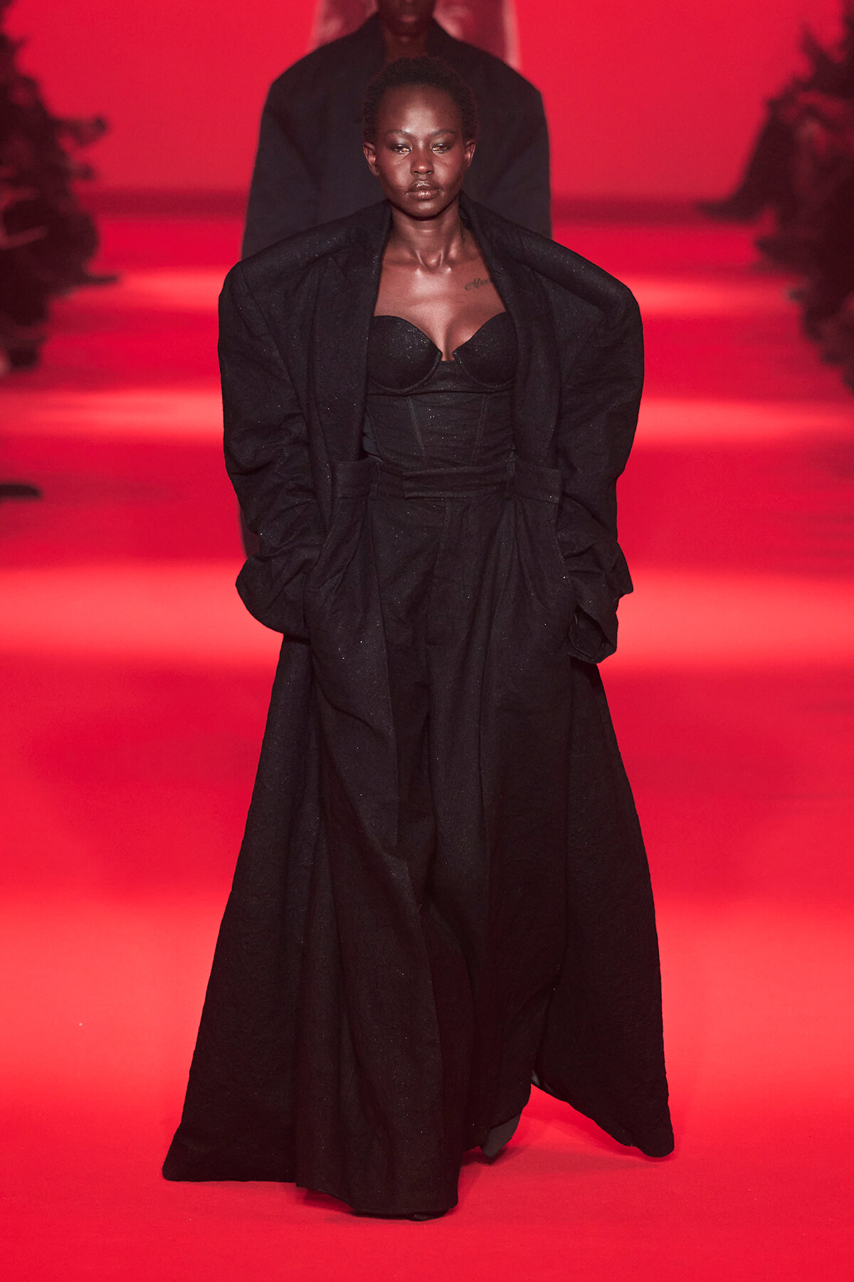 Female model walking a fashion show runway in a black corset top and long black coat against a red-lit backdrop.