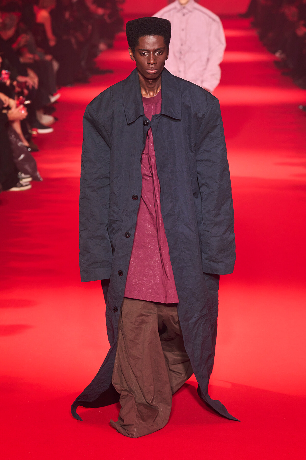 Model walking a red-lit runway in a long navy coat over a maroon top and brown loose pants.