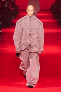 Male model walking a red runway in oversized pink check-pattern outfit with crinkled fabric and long sleeves.