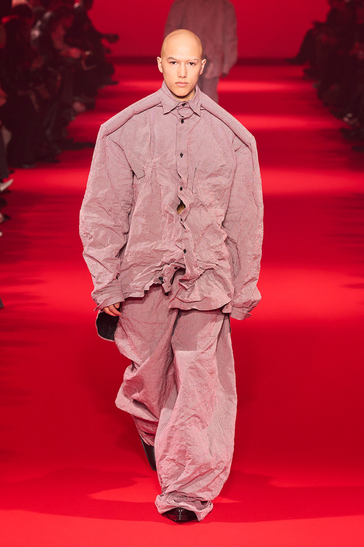 Male model walking a red runway in oversized pink check-pattern outfit with crinkled fabric and long sleeves.