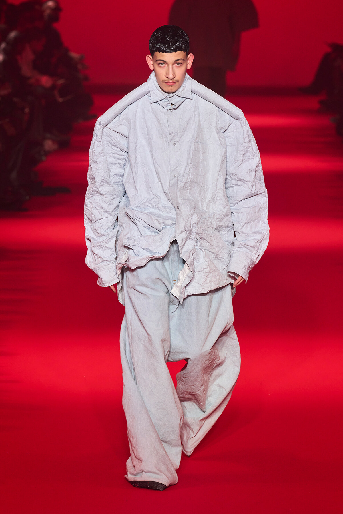 Male model walks a red runway in an oversized crumpled light-gray outfit with a high collar and loose, wrinkled fabric.
