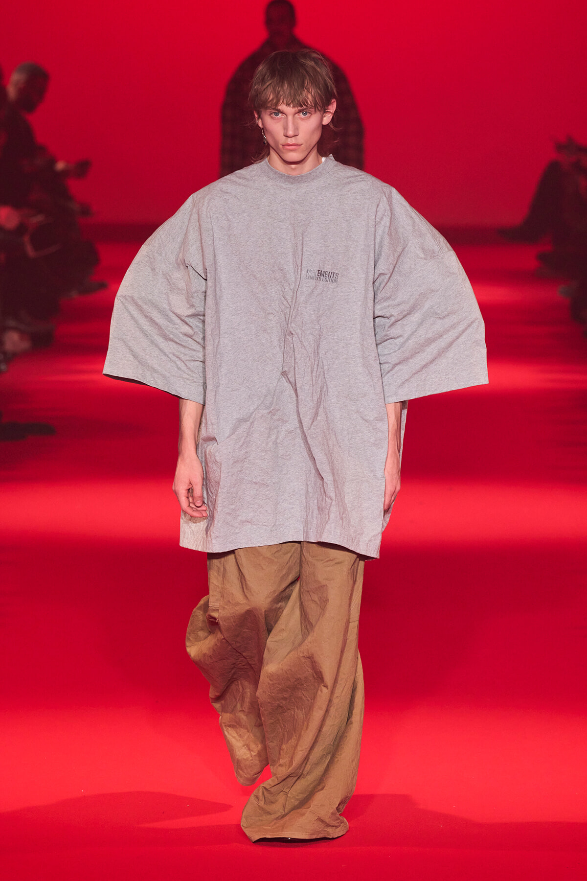 Model walks a runway in an oversized light gray tunic with wide sleeves and beige crinkled pants against a red backdrop, fashion show setting.