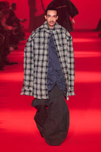 Male model walking a red-lit runway in an oversized black-and-white plaid coat over a dark blue shirt.