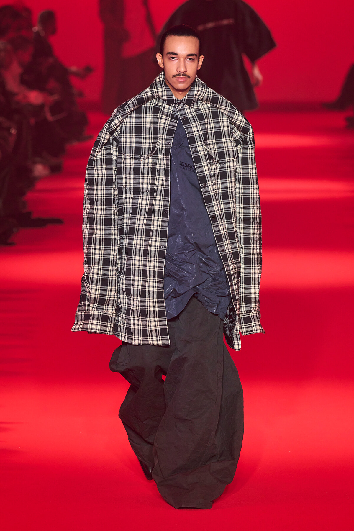 Male model walking a red-lit runway in an oversized black-and-white plaid coat over a dark blue shirt.