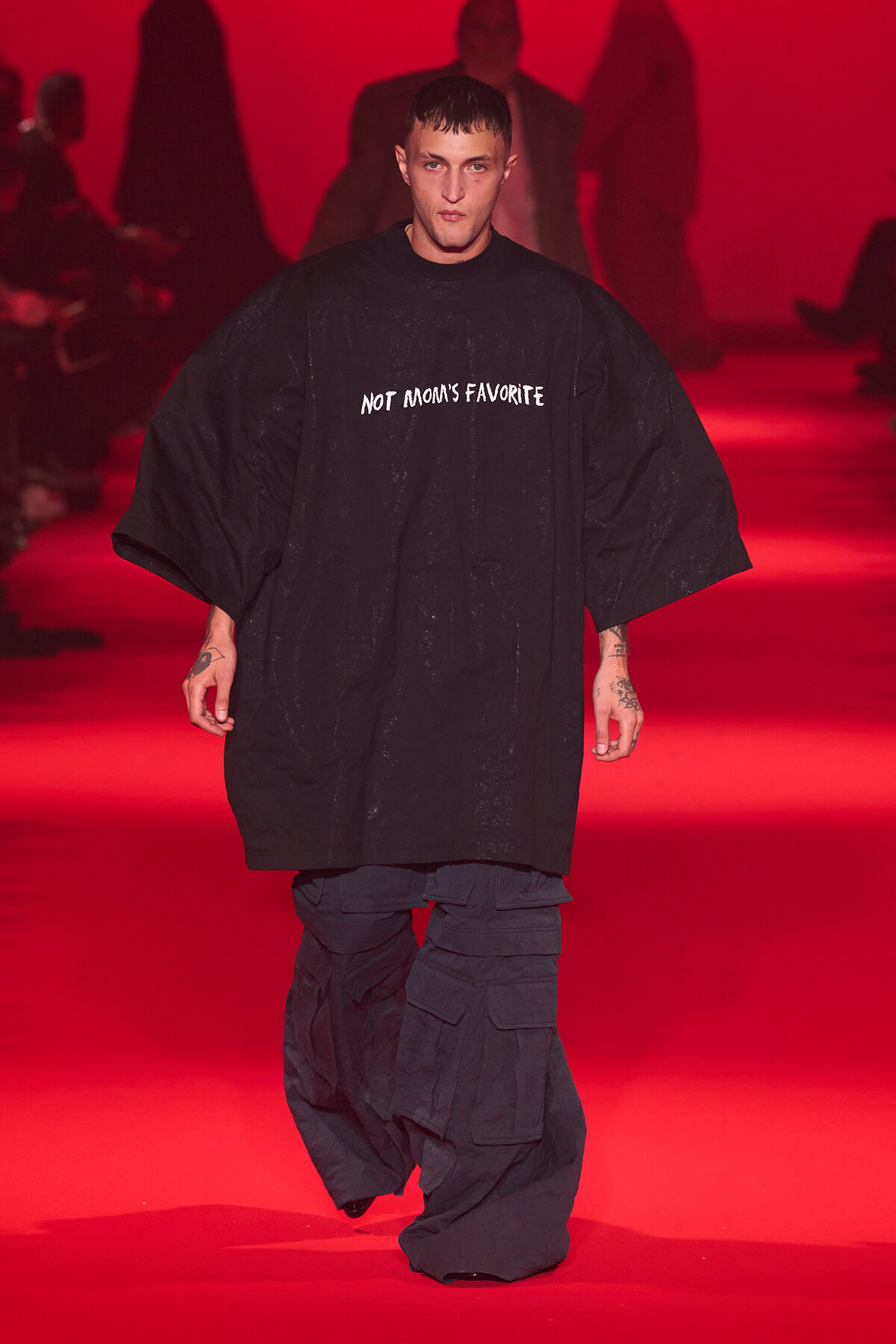 Male model walking a fashion runway in a loose black garment with 'NOT MOM'S FAVORITE' printed on it, red backstage lighting in the background