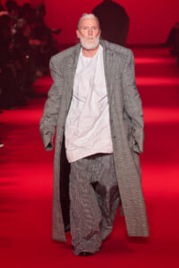 Older man with white beard wearing an oversized gray plaid suit walks the red-lit runway on a fashion show.