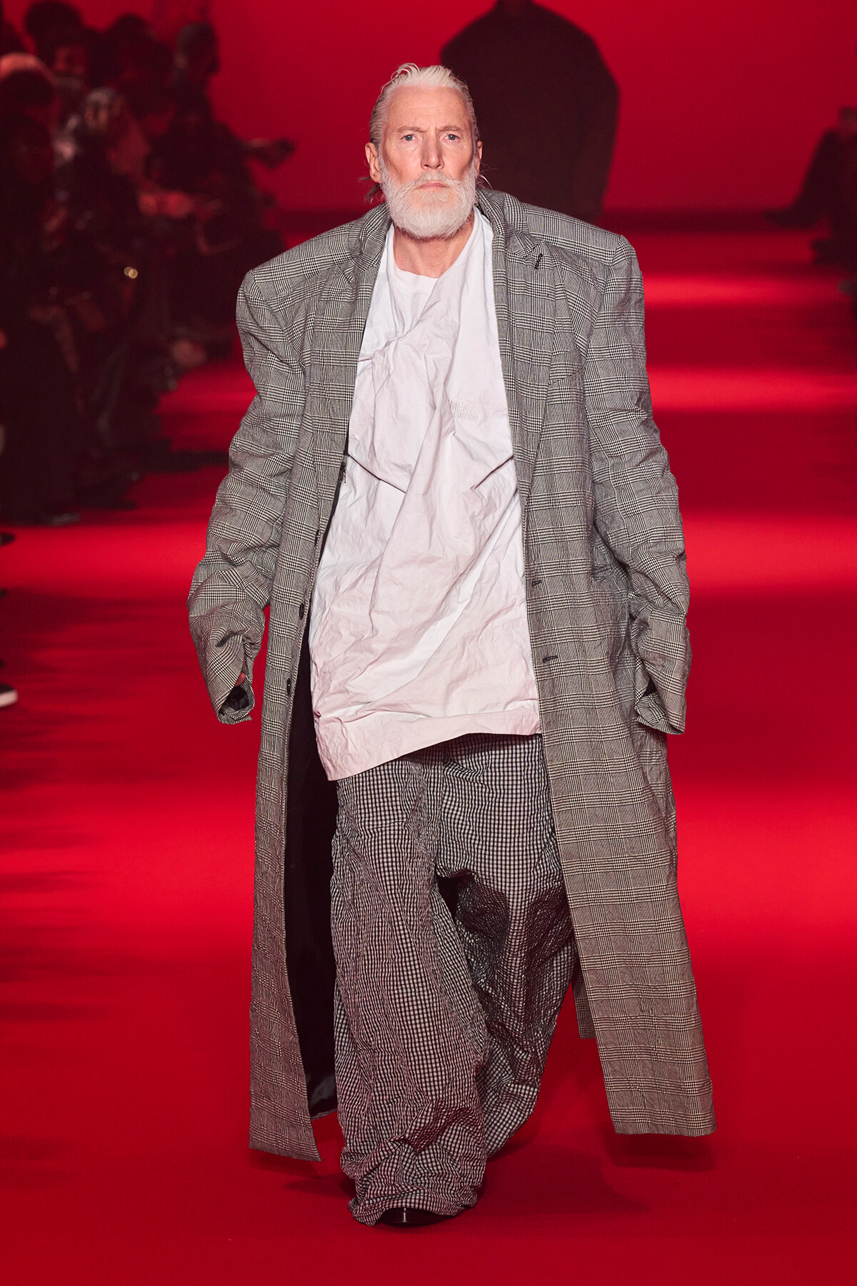 Older man with white beard wearing an oversized gray plaid suit walks the red-lit runway on a fashion show.