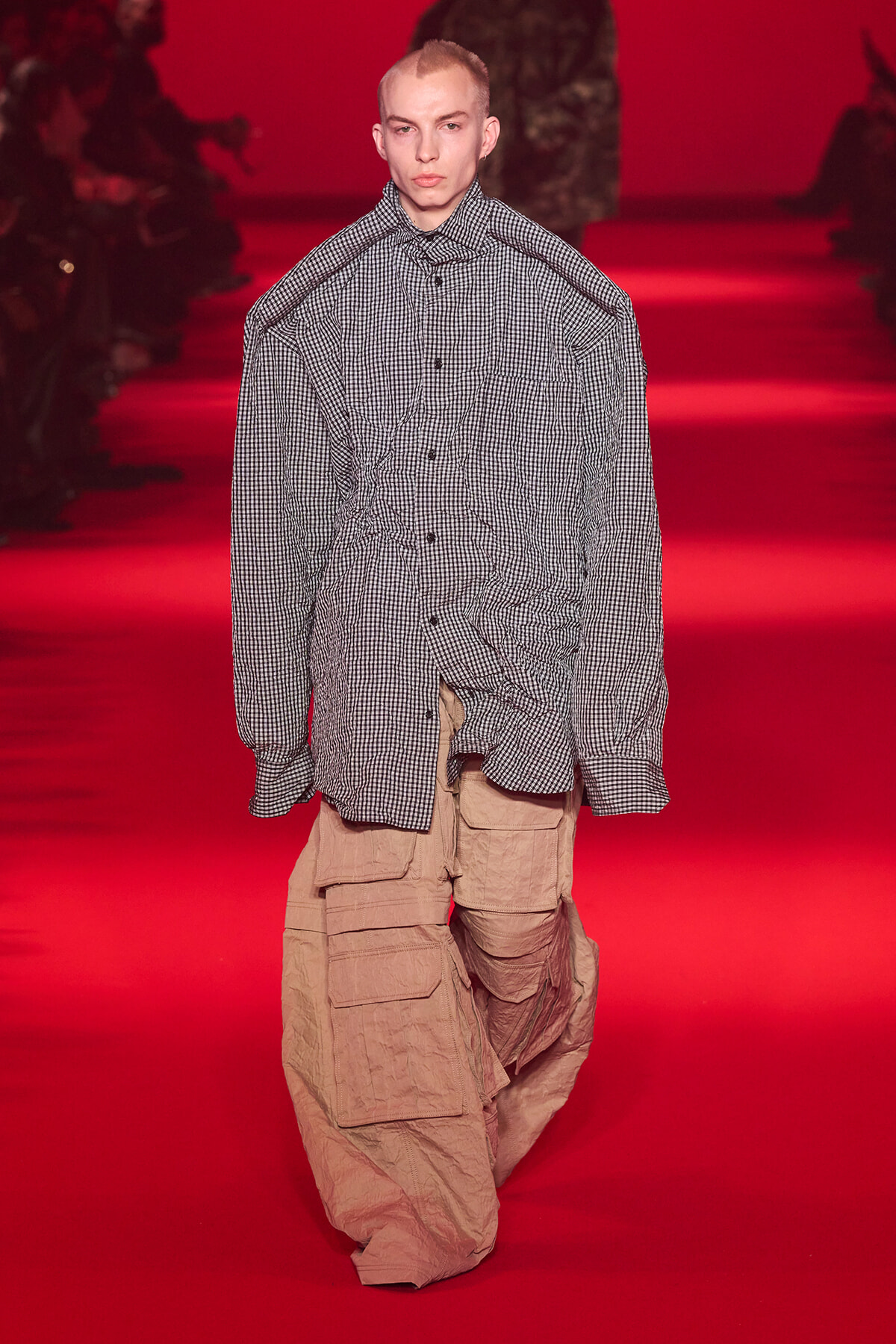 Model walks a fashion show wearing an oversized black-and-white checkered shirt and tan cargo pants under red runway lighting.