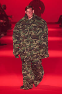 Model wearing camouflage military-style jacket and pants walking on a red-lit runway at a fashion show