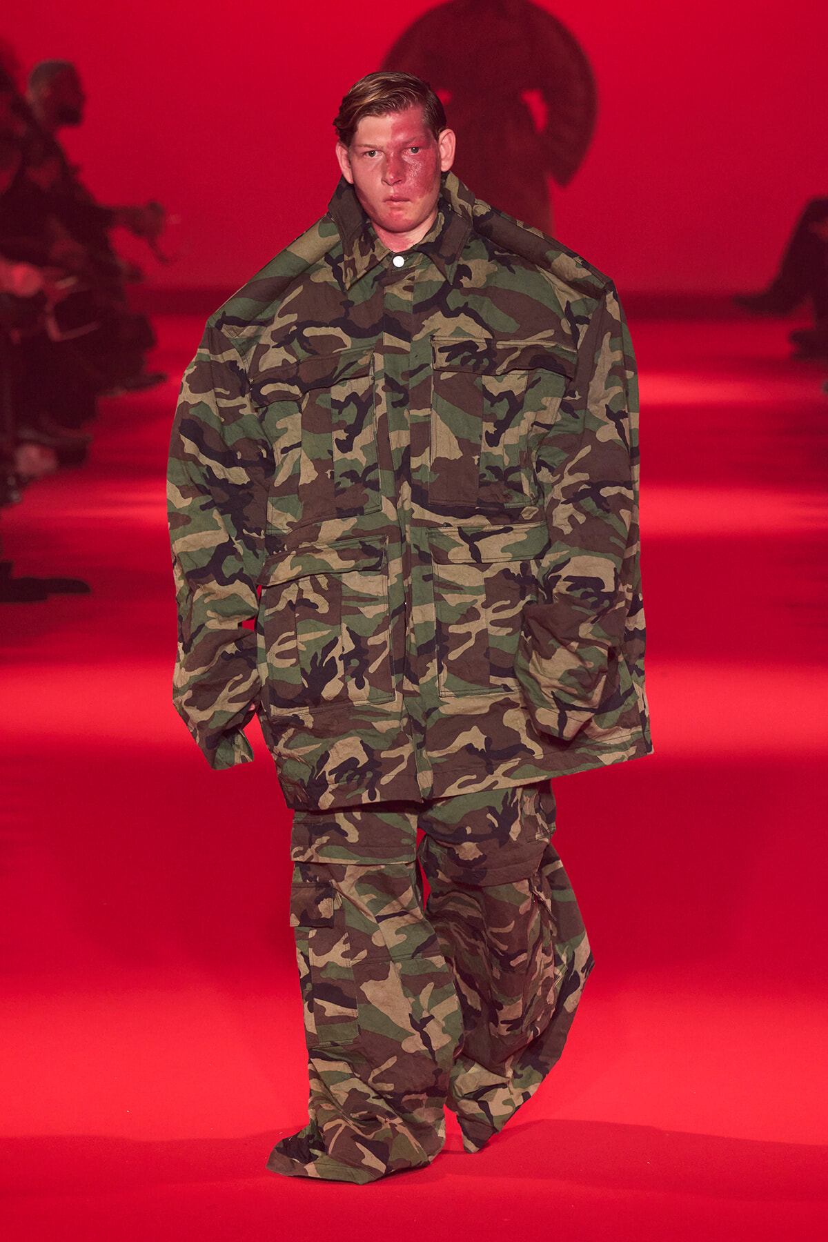 Model wearing camouflage military-style jacket and pants walking on a red-lit runway at a fashion show