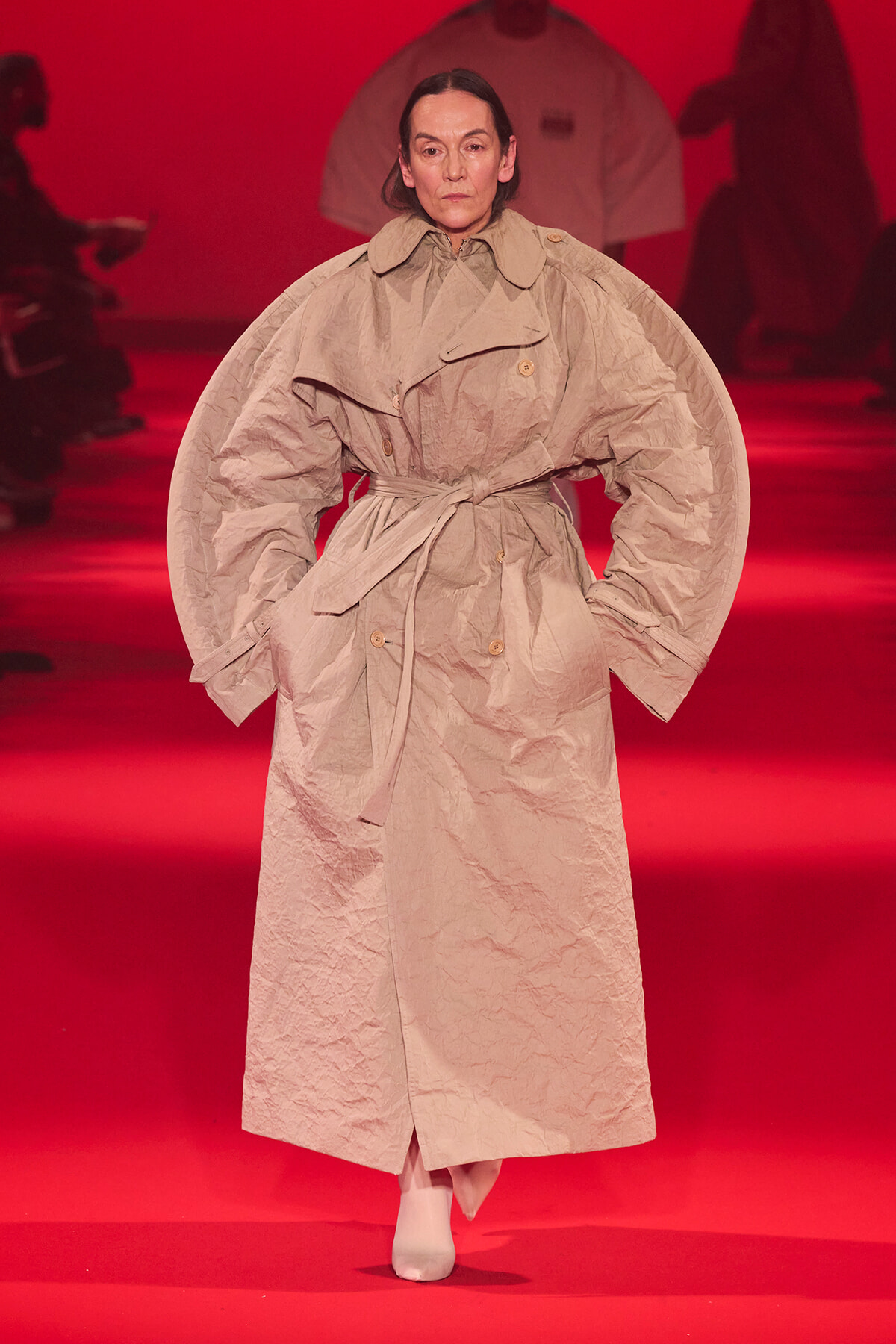 Model wearing a beige oversized trench with dramatic puff sleeves and belt on a red runway stage