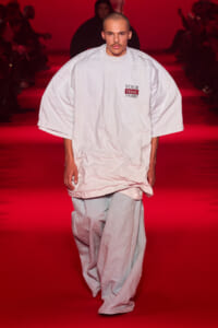 Male model walks a red-lit runway in an oversized white T-shirt with a small 'YOUR Basic T-SHIRT' logo on the chest.