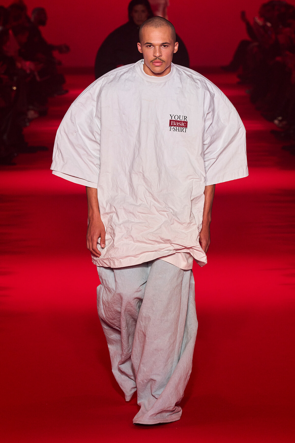 Male model walks a red-lit runway in an oversized white T-shirt with a small 'YOUR Basic T-SHIRT' logo on the chest.