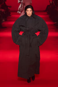 Model on a runway wearing an oversized black belted coat against a red-lit backdrop.
