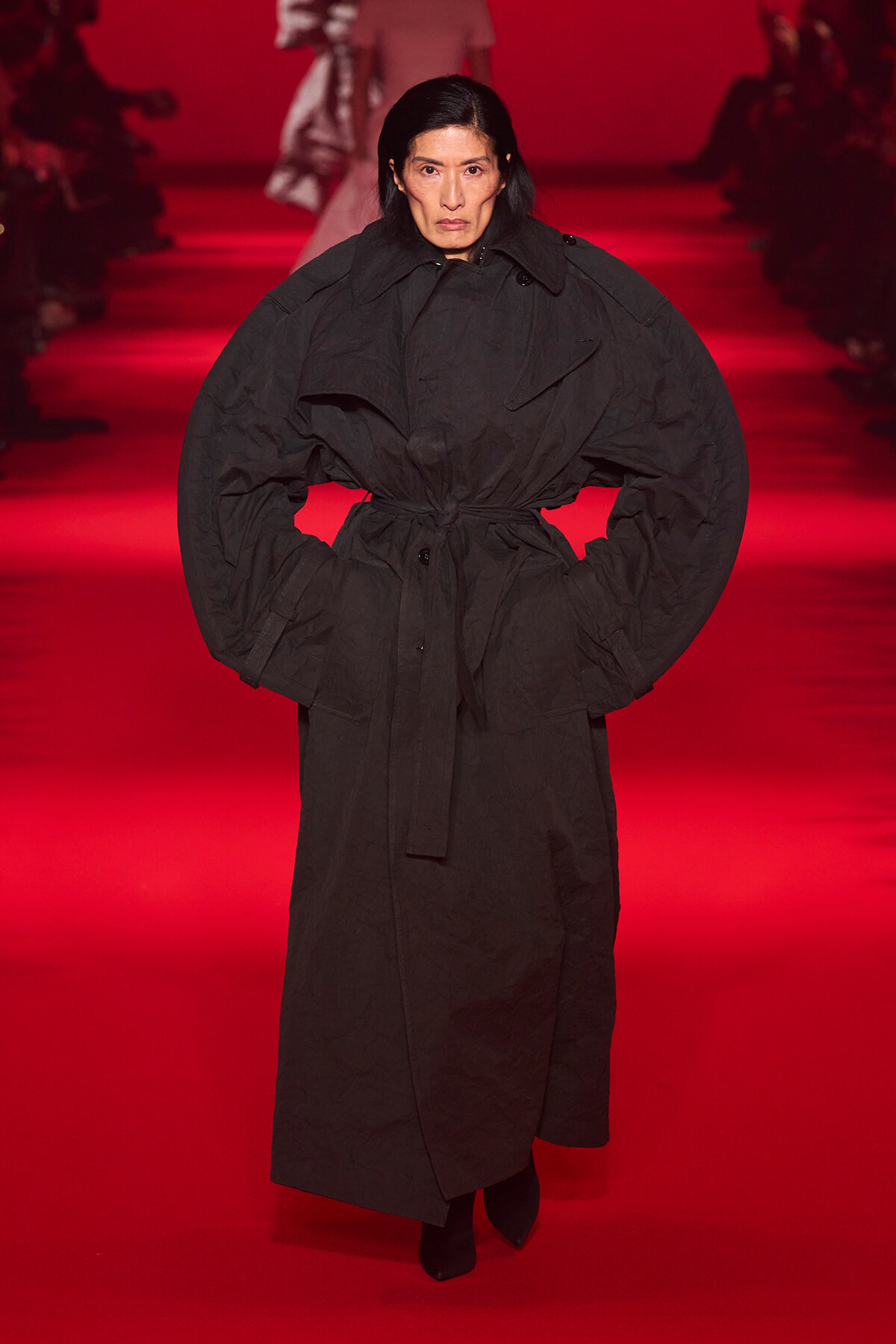 Model on a runway wearing an oversized black belted coat against a red-lit backdrop.