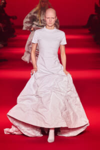 Bald model walking a red runway in a long white crinkled gown.