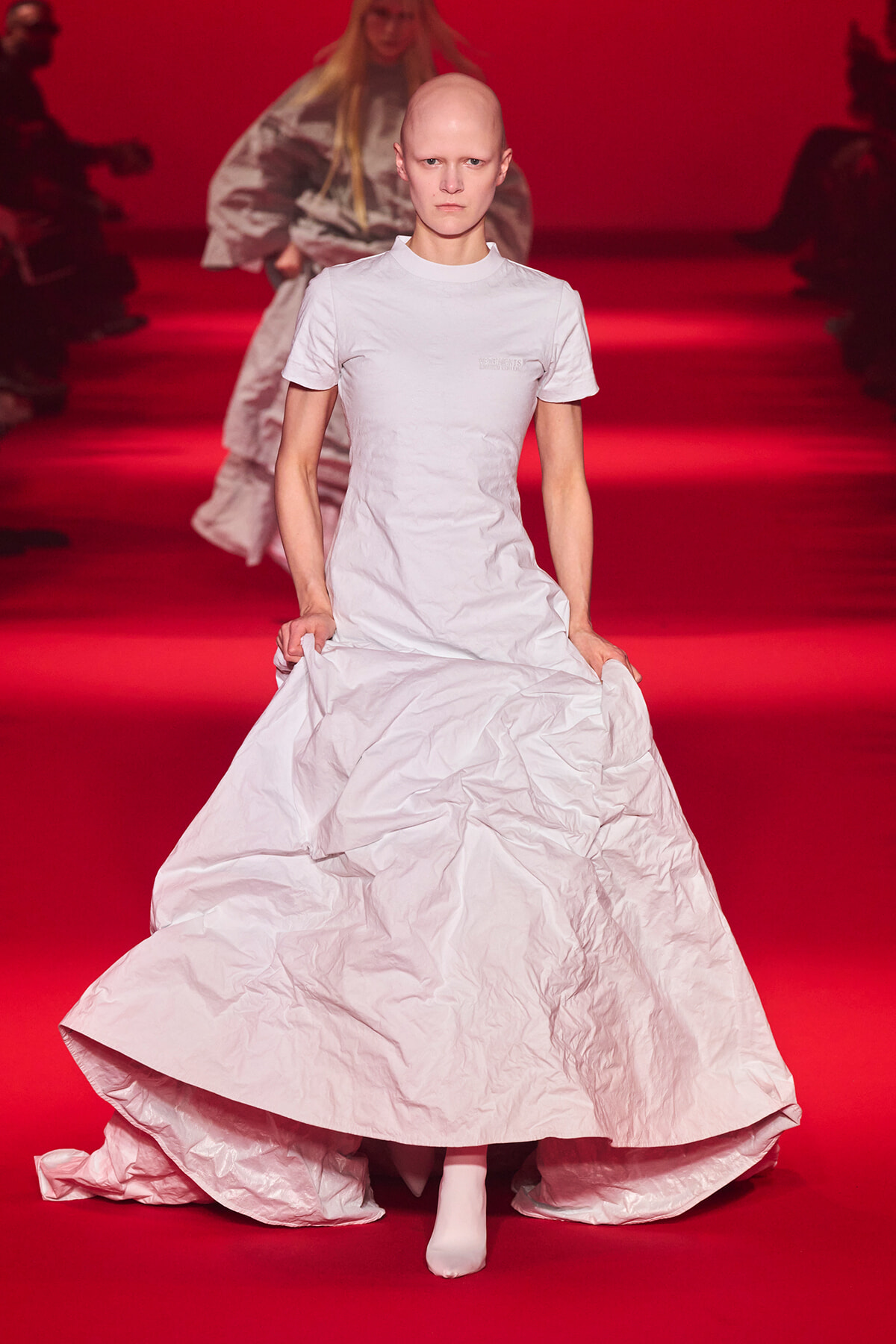 Bald model walking a red runway in a long white crinkled gown.