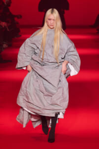 Model walking a runway in a voluminous crinkled gray dress with long blonde hair under red lighting theater backdrop.