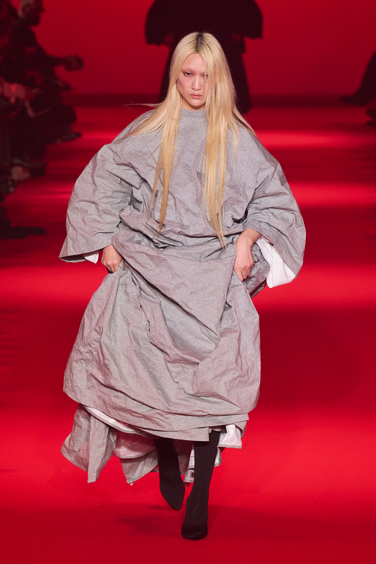 Model walking a runway in a voluminous crinkled gray dress with long blonde hair under red lighting theater backdrop.