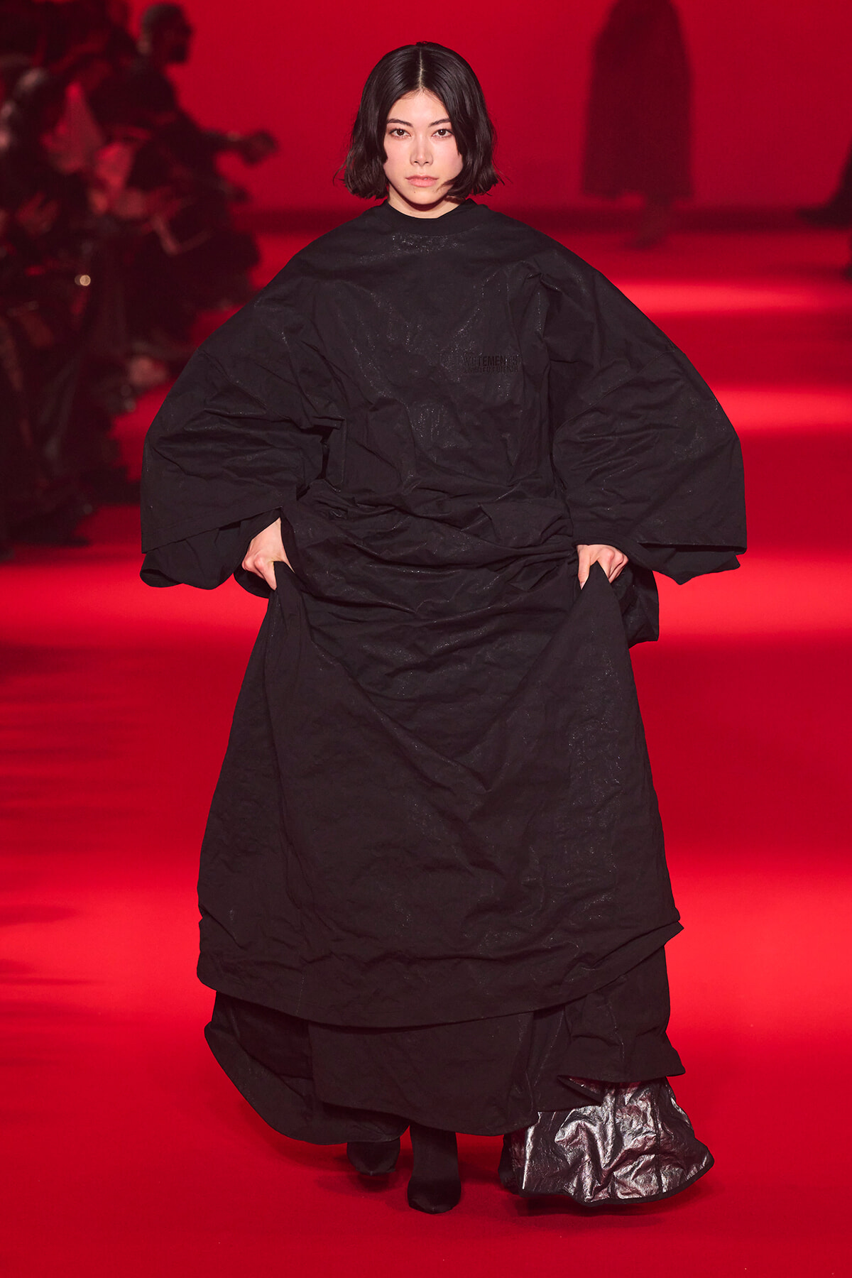 Model wearing an oversized black cape-like garment on a red-lit runway.