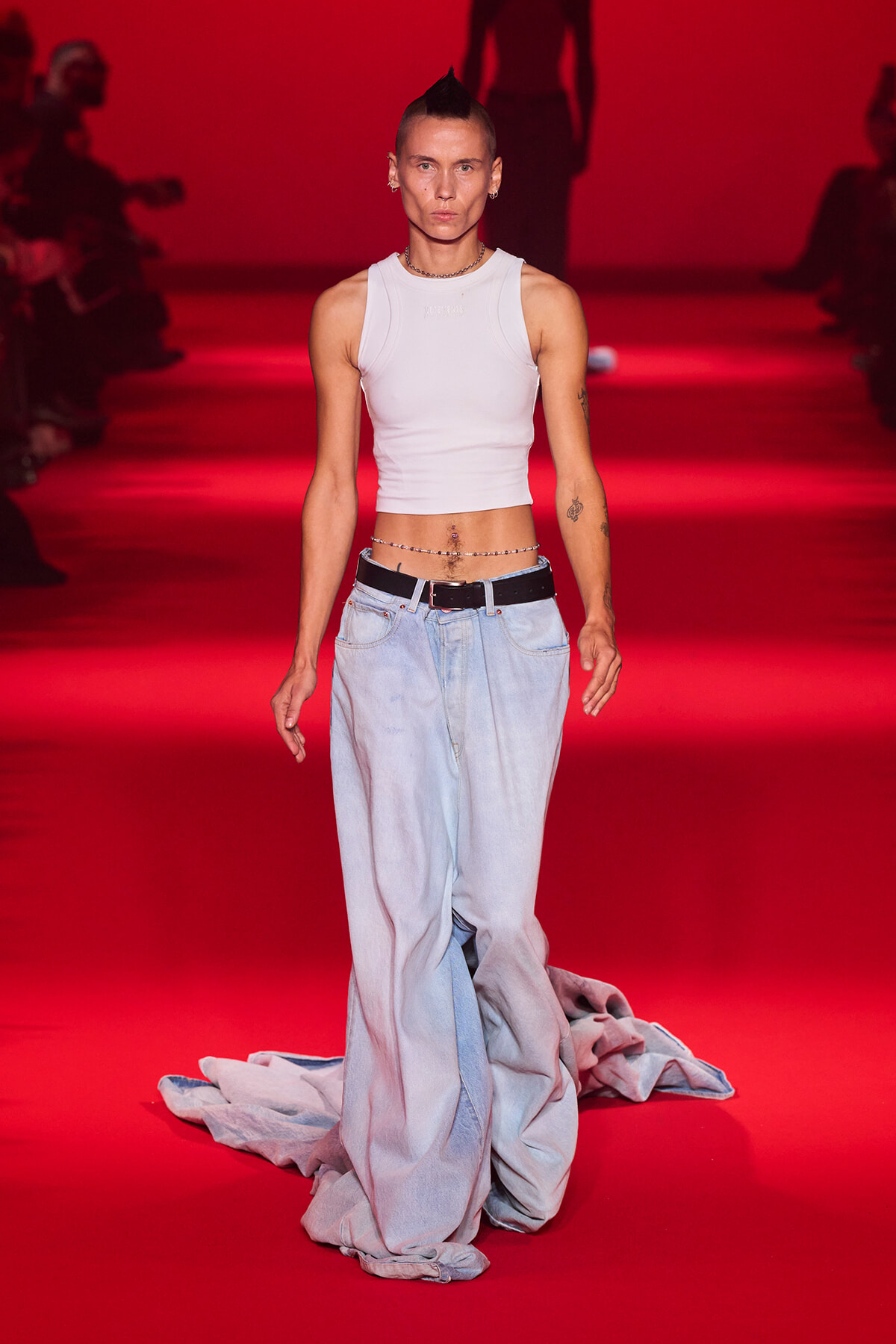 Model walking a red-lit runway in a white cropped tank top and oversized light-wash jeans, accessorized with a belt and necklace.