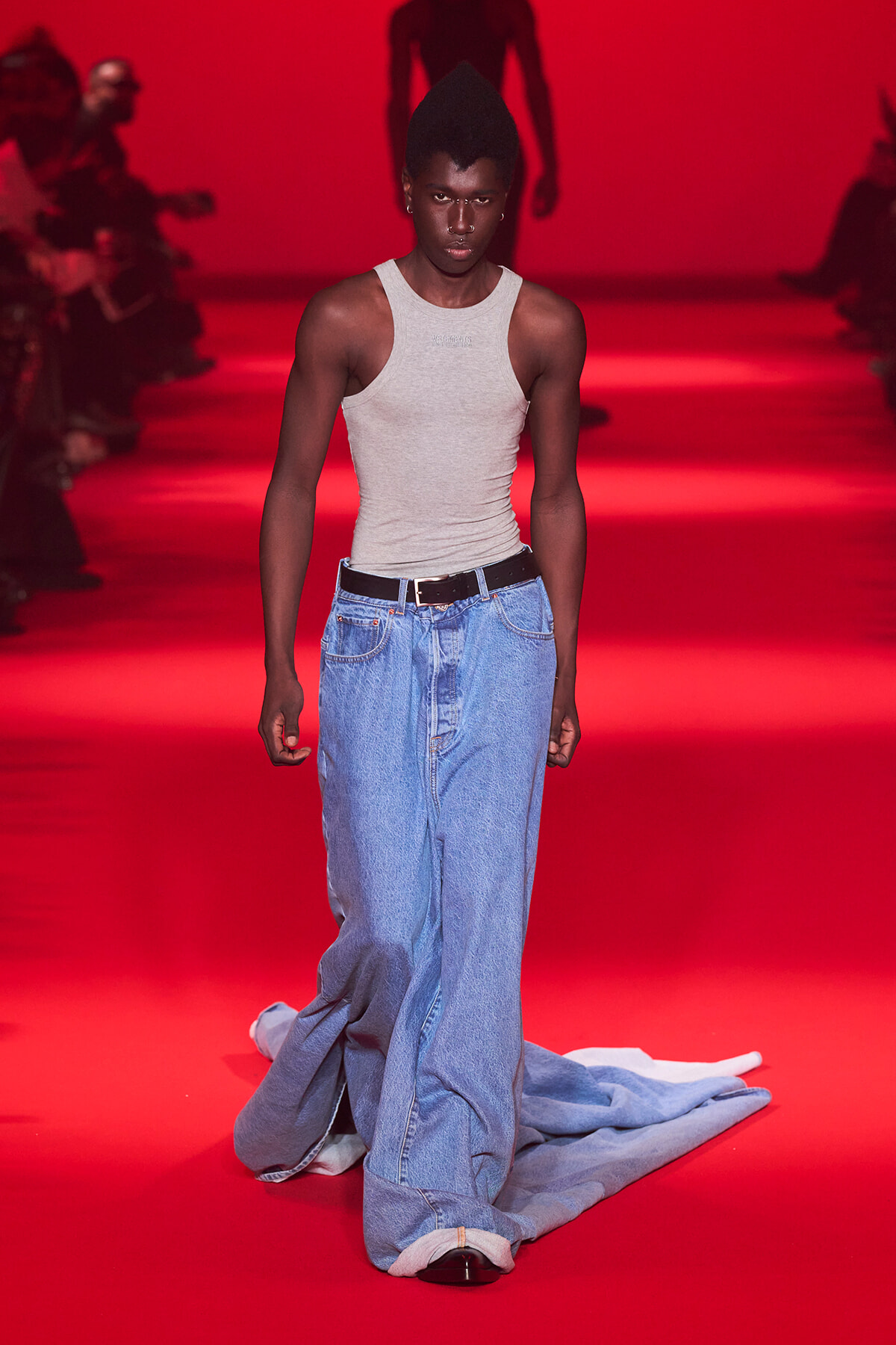 Male model walking a red-lit runway in a gray sleeveless top and oversized blue jeans with a black belt.