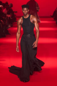 Male model walks a red-lit runway in a black tank top and black wide-leg pants, facing the camera with a confident pose.