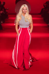 Model walking a runway in a gray sleeveless top and bright red striped wide-leg pants under red lighting, hands in pockets.