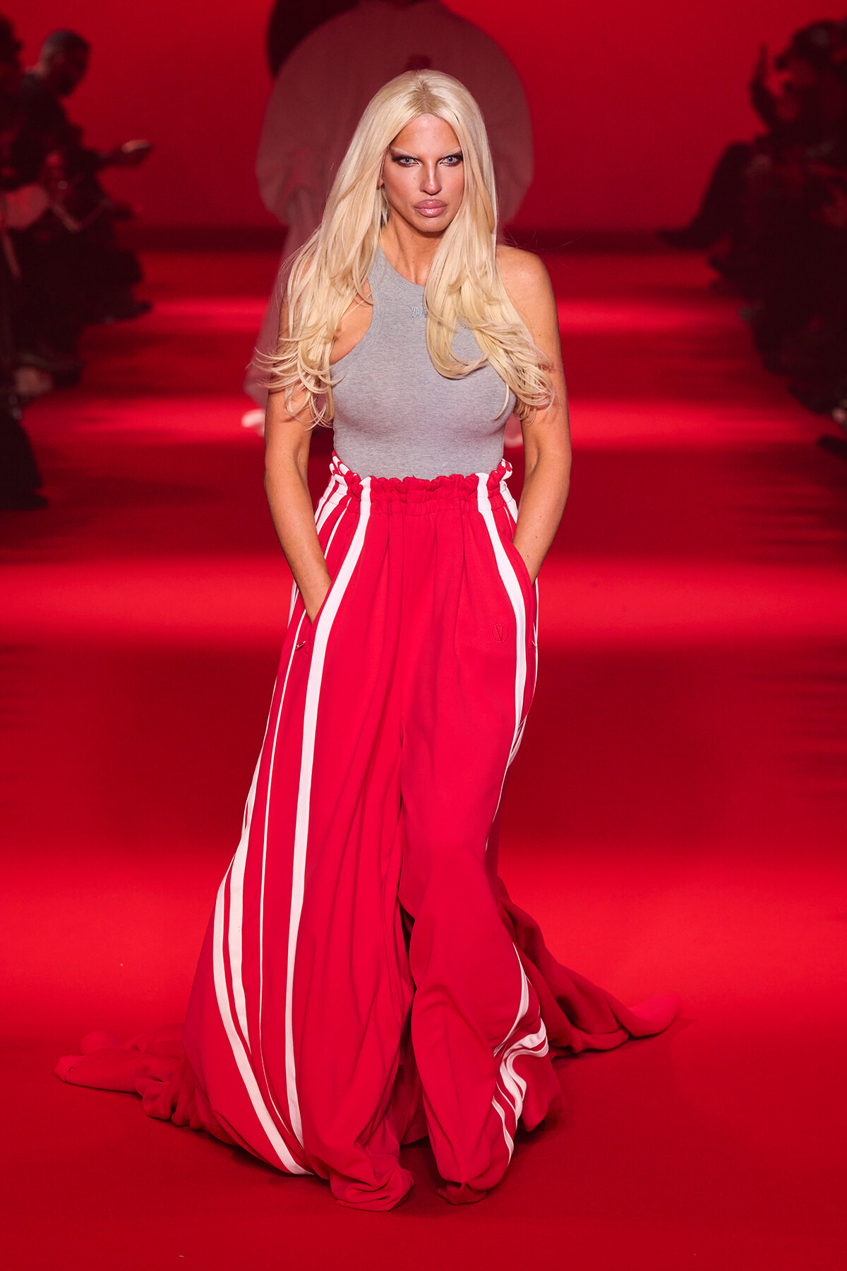 Model walking a runway in a gray sleeveless top and bright red striped wide-leg pants under red lighting, hands in pockets.