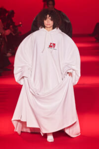 Model walks a runway in a voluminous white hooded garment with an 'I'M SHY' logo on the chest, red lighting in the background.