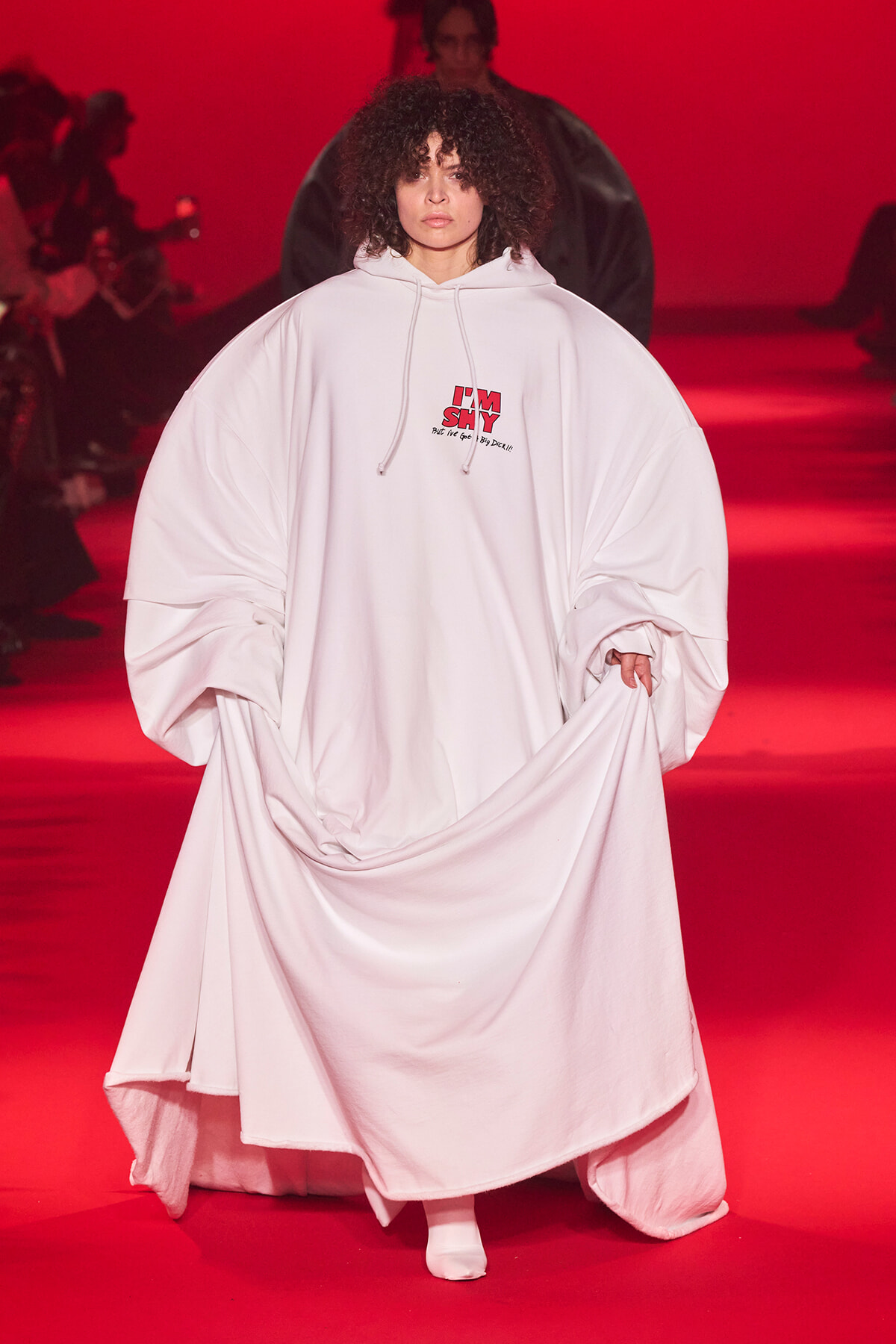 Model walks a runway in a voluminous white hooded garment with an 'I'M SHY' logo on the chest, red lighting in the background.
