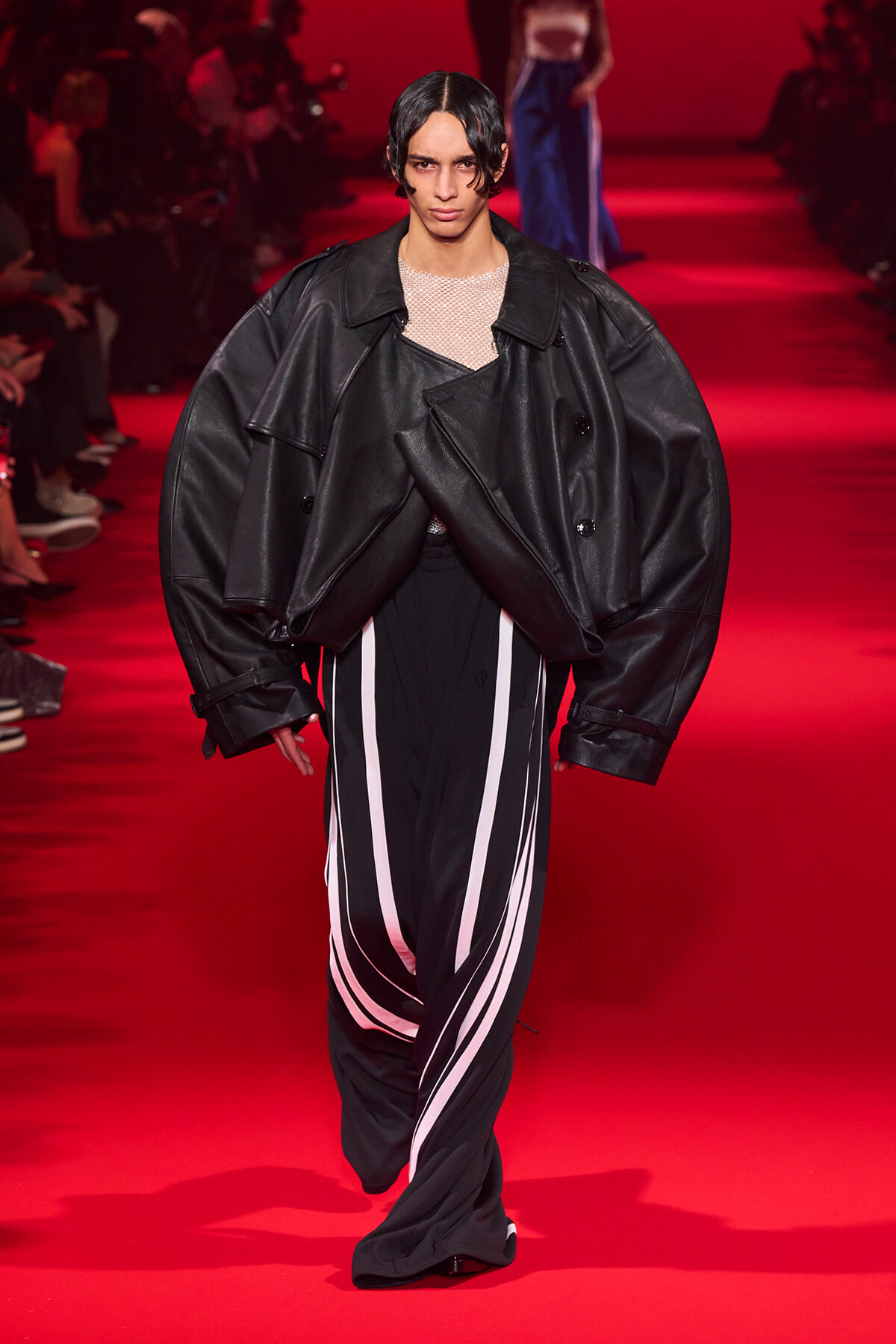 Model walks a red runway wearing an oversized black leather jacket layered over a sheer mesh top and black pants with white curved stripes.