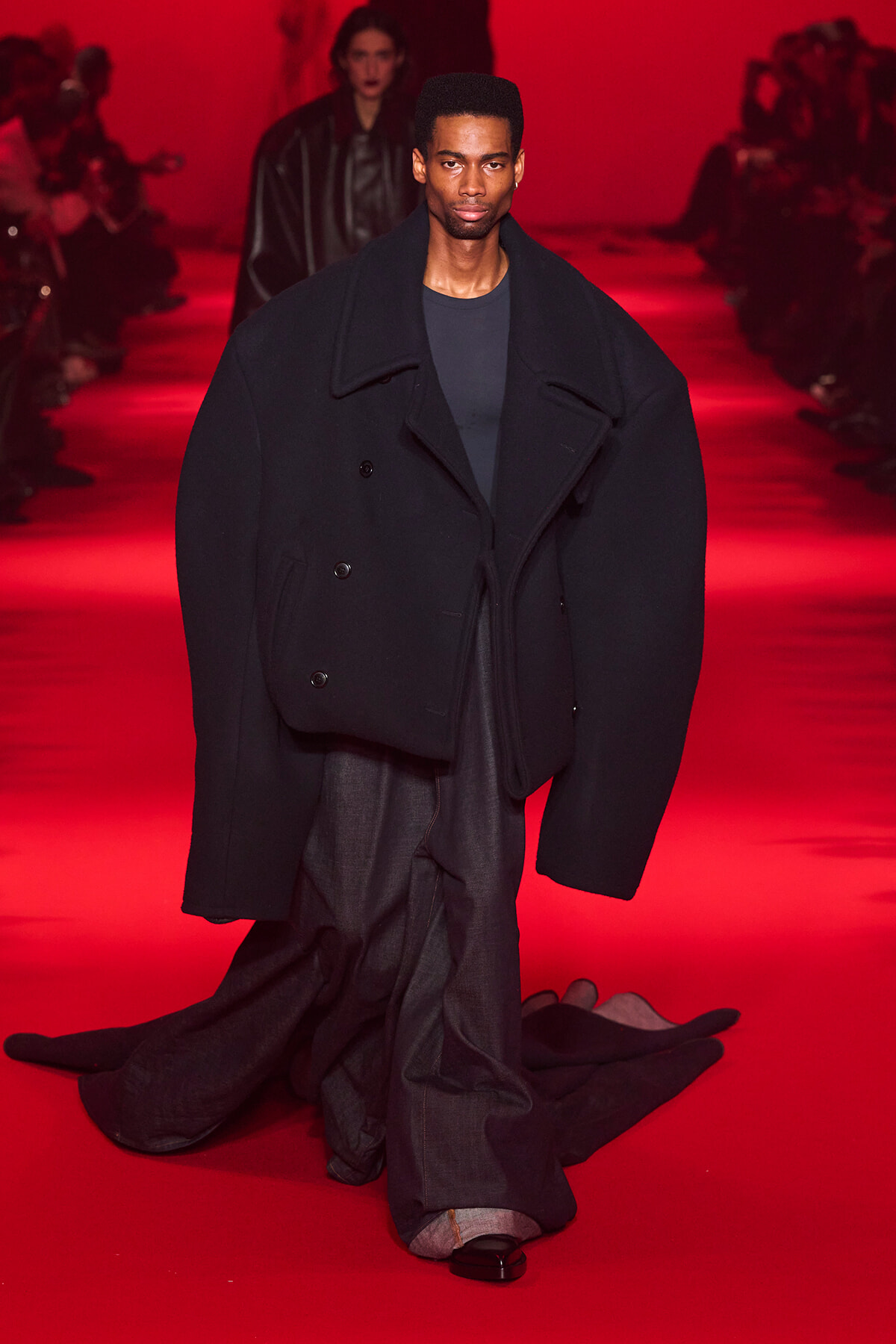 Male model wearing an oversized black coat on a red-lit runway.