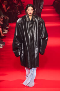 Model walking a fashion show in an oversized black leather coat on a red-lit runway.