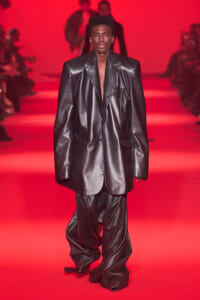 Model walks the runway in an oversized black leather suit against a red backdrop, bold shoulders and long jacket tails visible.