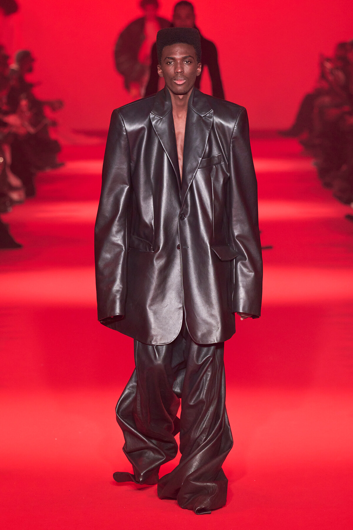 Model walks the runway in an oversized black leather suit against a red backdrop, bold shoulders and long jacket tails visible.