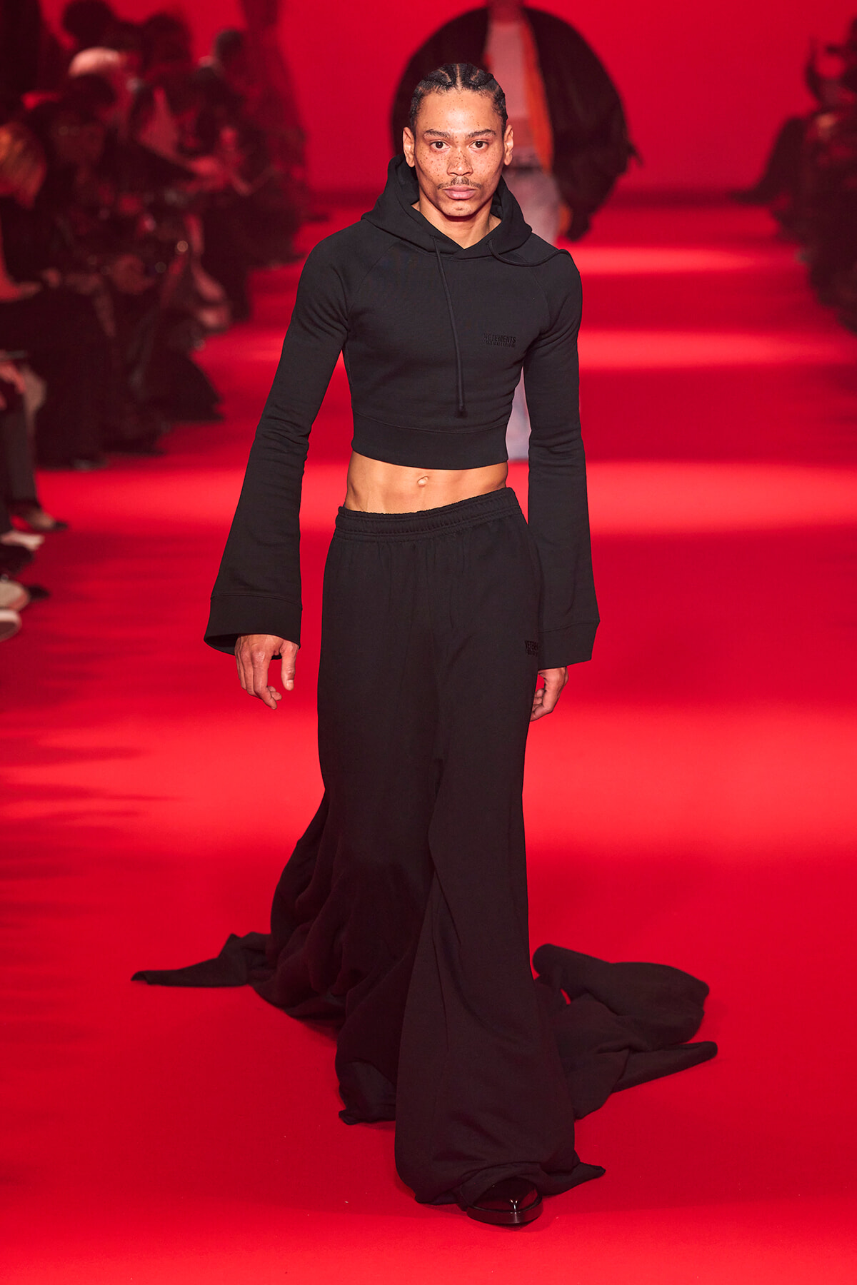 Male model walks the red-lit runway in a black cropped hoodie and wide-leg pants, center frame.