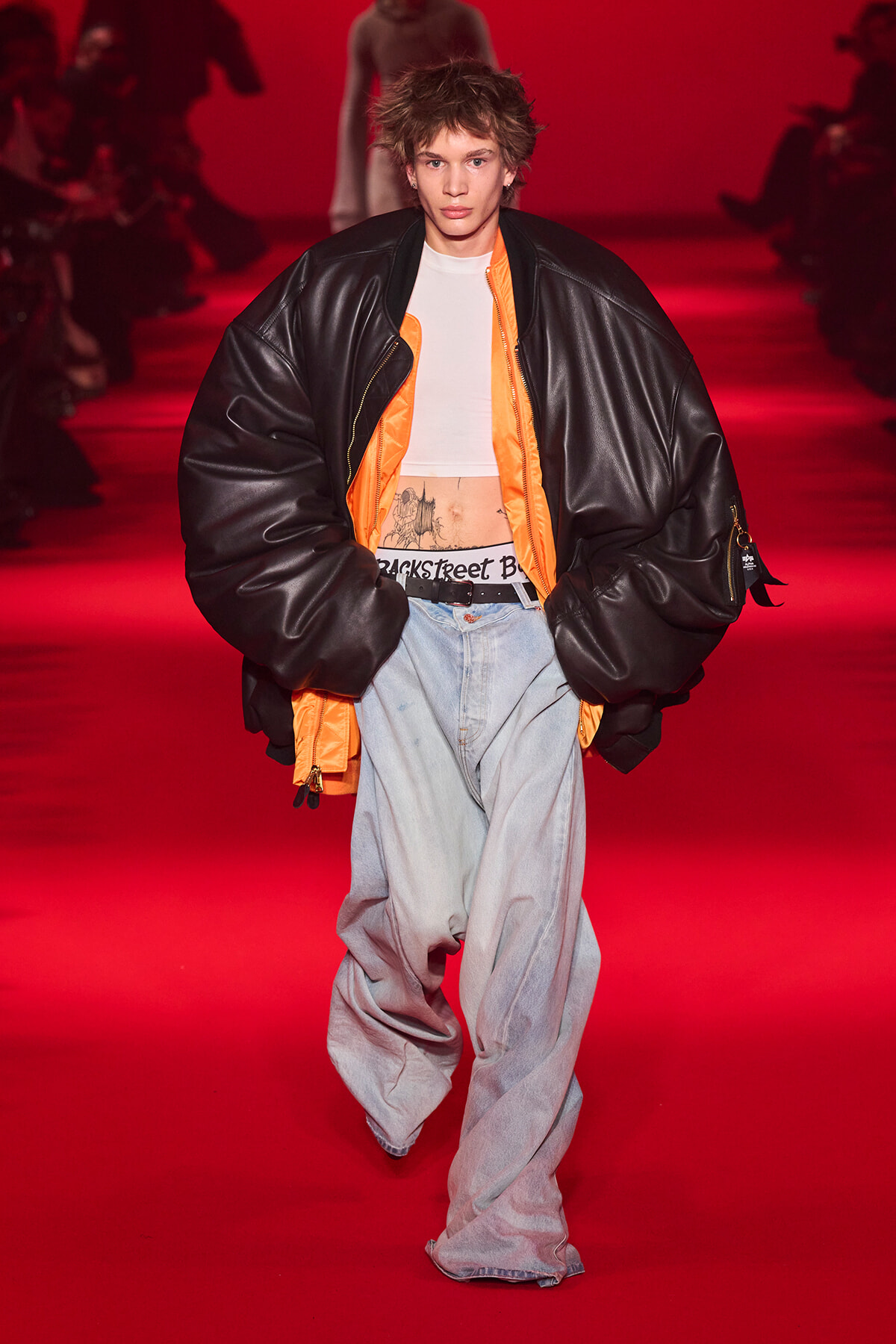 Runway model in an oversized black puffer jacket with orange lining over a white crop top and baggy light-wash jeans, against a red backdrop with seated audience in the background.