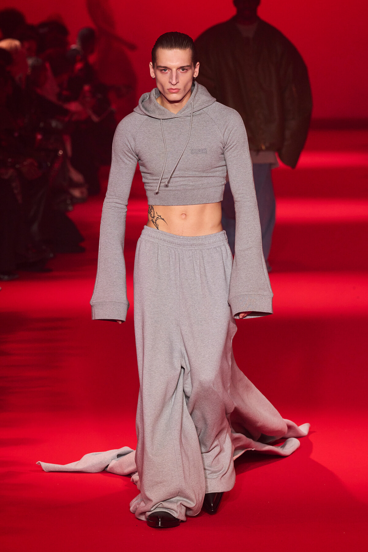 Male model walks a red-lit runway in a gray cropped hoodie and loose gray sweatpants, tattoo visible on his left abdomen.
