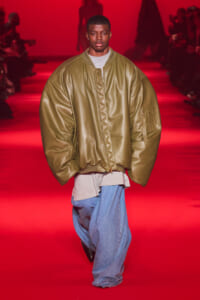 Male model walks a red-lit runway wearing an oversized olive bomber jacket, gray tee, and baggy blue jeans.