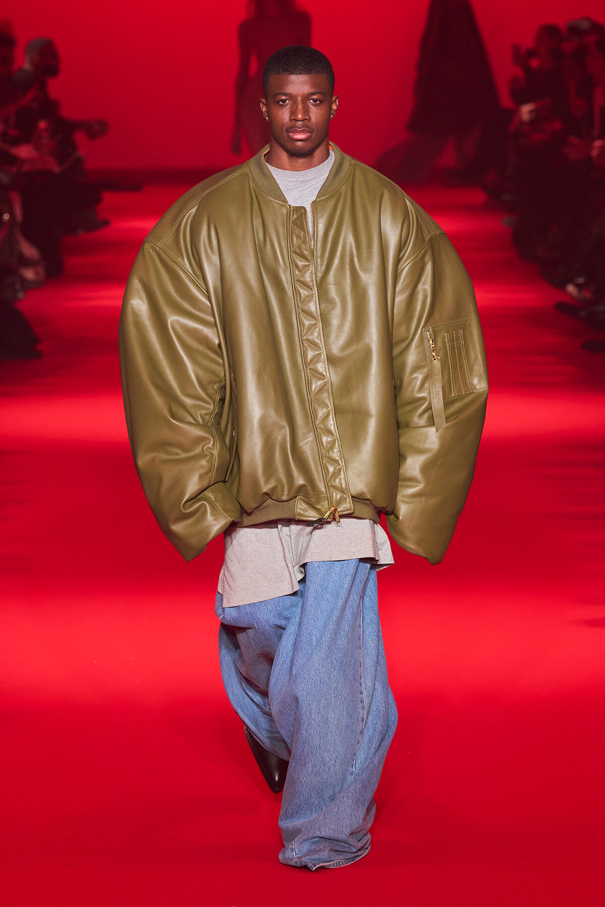 Male model walks a red-lit runway wearing an oversized olive bomber jacket, gray tee, and baggy blue jeans.