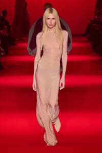 Model walking a red-carpet runway in a form-fitting pale pink dress under red lighting.