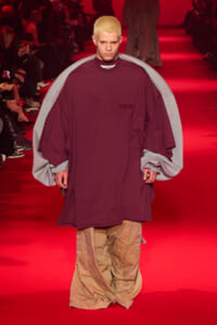 Male runway model wearing an oversized burgundy top with grey cape-like sleeves, walking on a red-lit catwalk; tan cargo pants visible at the bottom of the outfit.