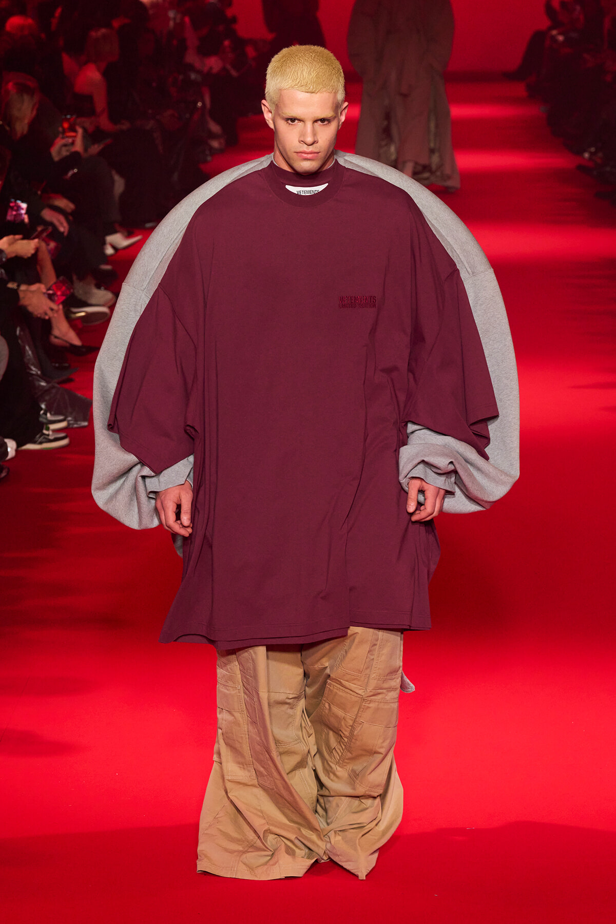 Male runway model wearing an oversized burgundy top with grey cape-like sleeves, walking on a red-lit catwalk; tan cargo pants visible at the bottom of the outfit.