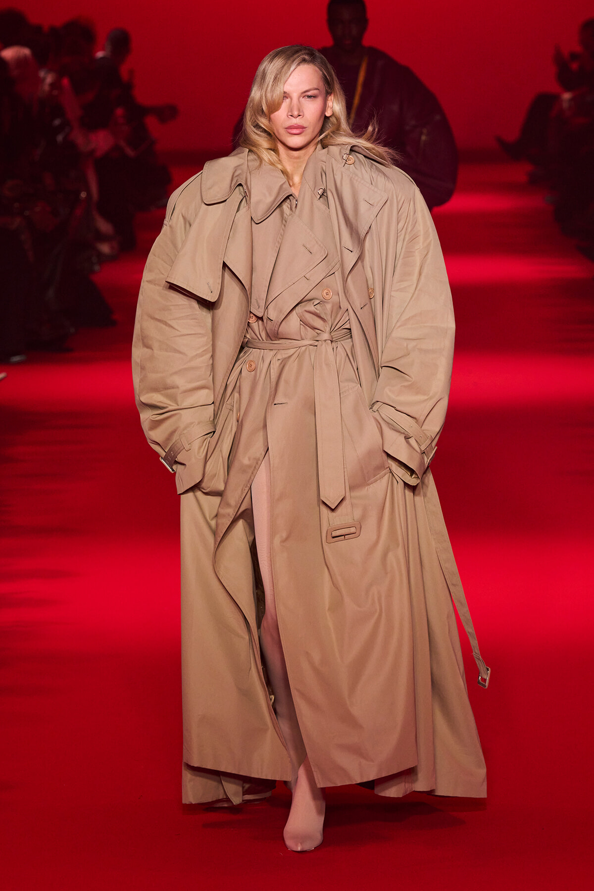 Model walking the runway in an oversized beige trench coat with belt on a red-lit catwalk.
